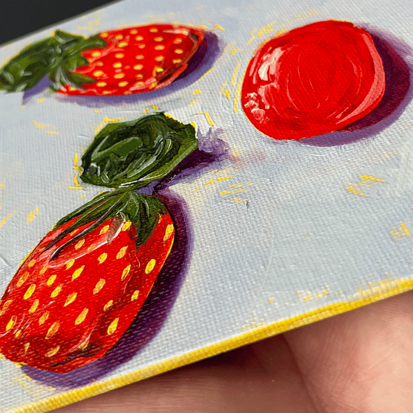 Close-up of a hand holding a small painting of strawberry candies on a textured surface.